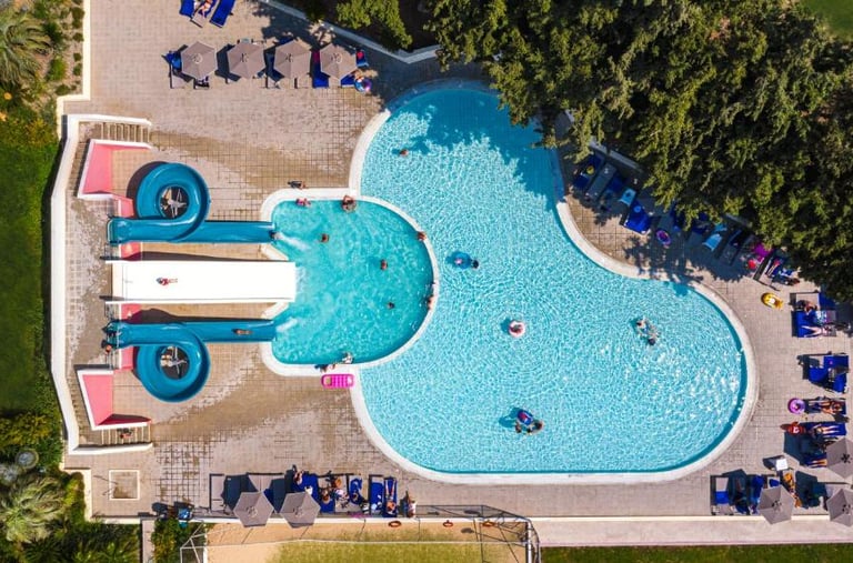 Aerial view of a luxury resort swimming pool with blue water slides and guests relaxing on lounge chairs.