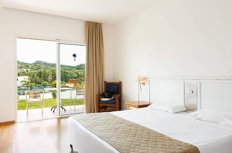 Bright modern hotel room with white bedding and a balcony overlooking a pool and hills.