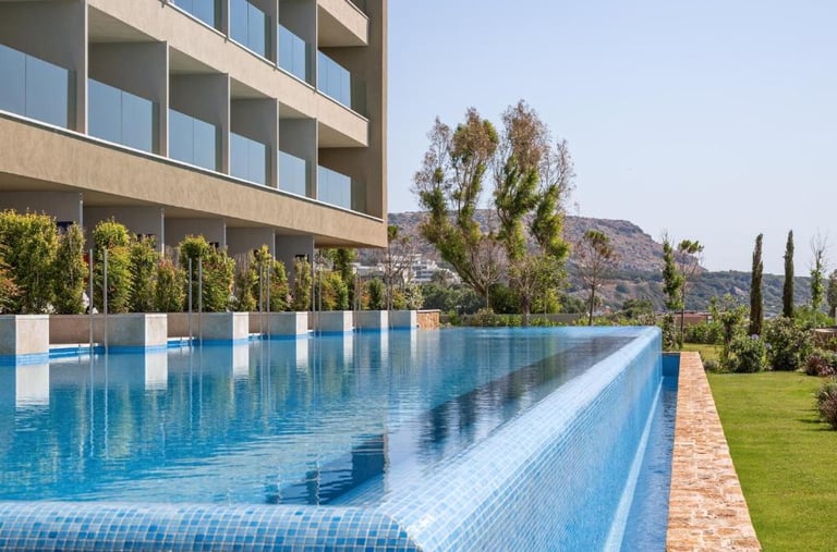 Luxury hotel infinity pool with blue tiles overlooking a lush garden and modern building balconies.
