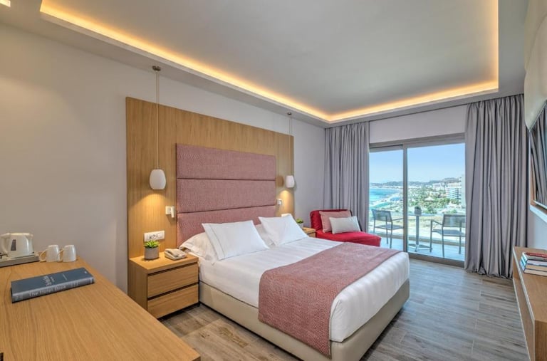 Modern hotel room with a double bed, pink headboard, and balcony overlooking the ocean.