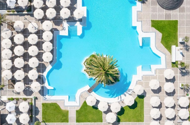Aerial view of a luxury resort swimming pool with white umbrellas and a central palm tree island.
