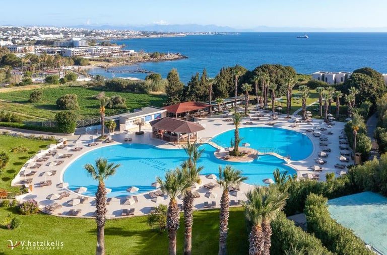 Aerial view of a luxury Greek island resort with a blue swimming pool and palm trees overlooking the Mediterranean Sea.