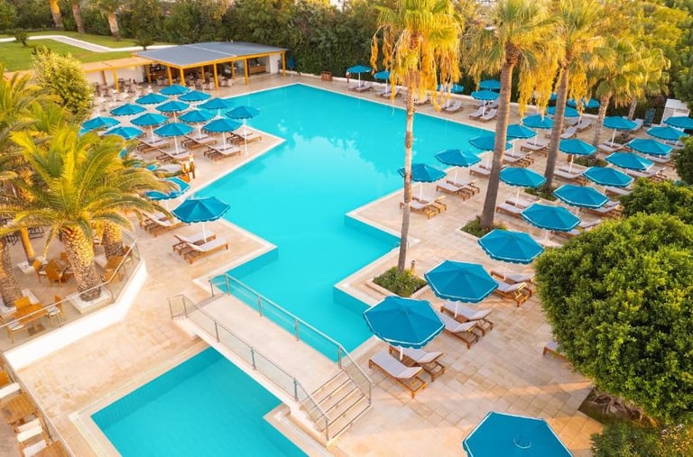 Aerial view of a luxury resort swimming pool with blue umbrellas, palm trees, and lounge chairs.