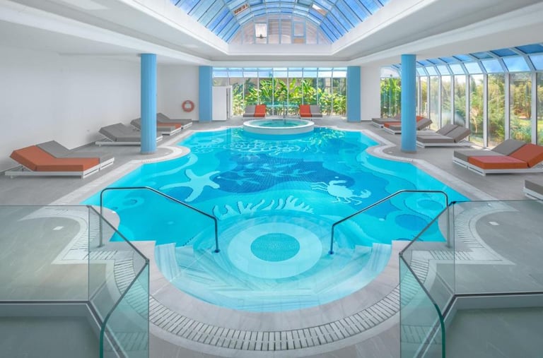 Luxury indoor swimming pool with sea creature mosaic tiles, glass skylight, and lounge chairs.