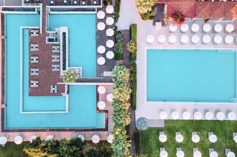 Aerial view of a luxury hotel resort featuring turquoise swimming pools, white sun umbrellas, and palm trees.