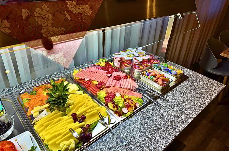 Hotel breakfast buffet featuring platters of sliced cheese, cold cuts, salmon, and yogurt containers.