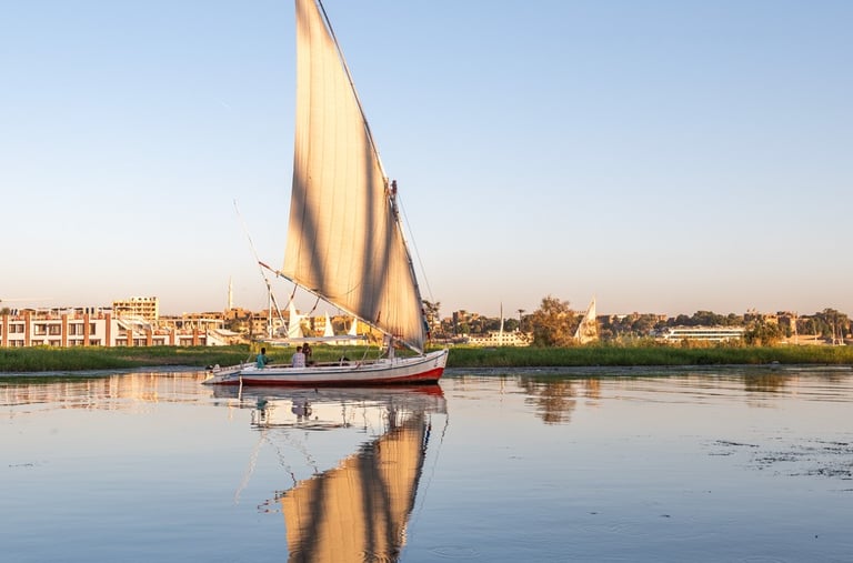 A traditional Egyptian felucca sailboat with a white sail reflecting on the calm Nile River at sunset.