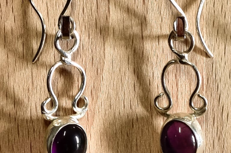 Handmade sterling silver drop earrings with oval purple amethyst gemstones on a wooden background.