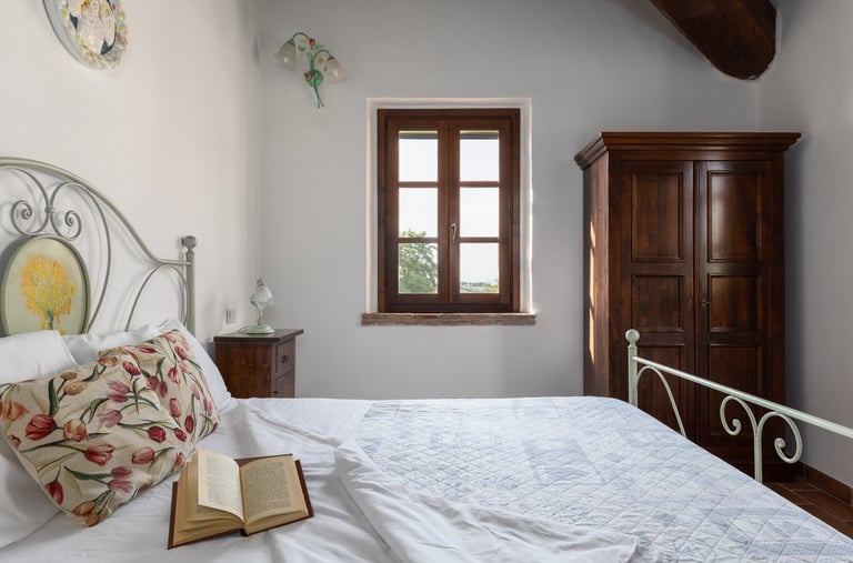 Rustic bedroom with a white iron bed frame, floral pillows, an open book, and a vintage wooden wardrobe.