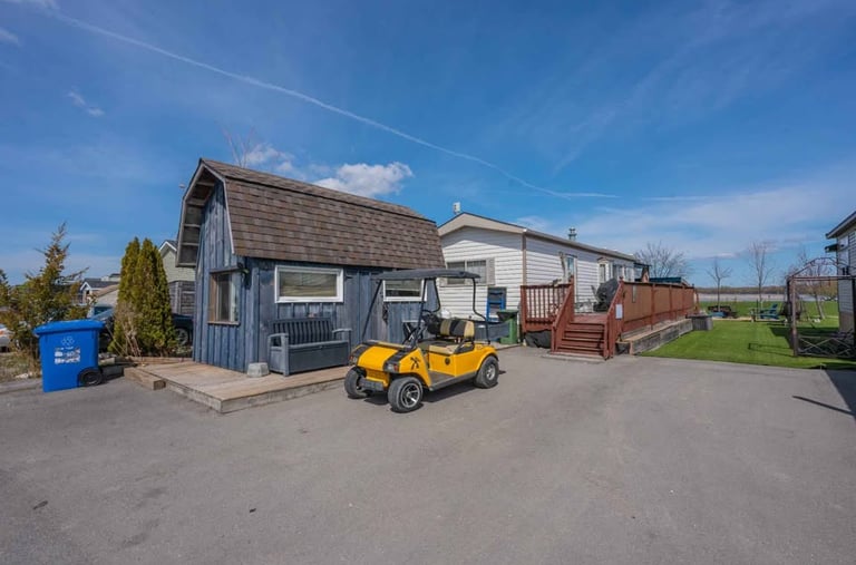 Yellow golf cart parked outside a blue shed and mobile home with wooden deck under a blue sky.