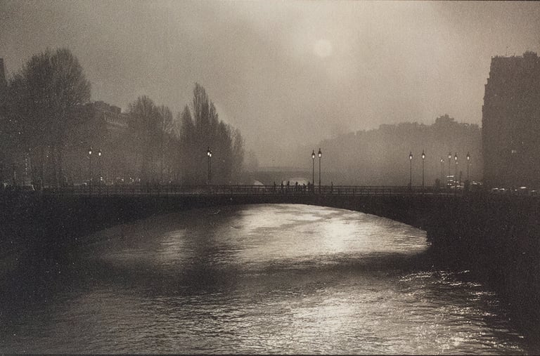 Platinum and silver photography of a foggy bridge over a river in Paris with soft city lights.