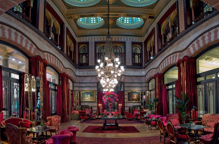 Elegant hotel lounge with ornate stained glass domes, a crystal chandelier, and red velvet armchairs.