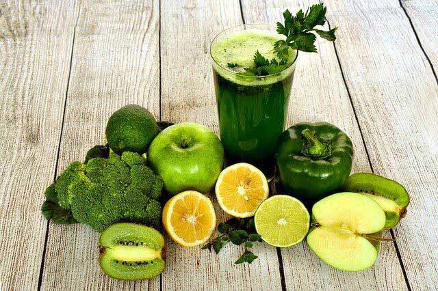 Copo de suco verde sobre mesa de madeira, acompanhado de maçã verde, limão, kiwi, pimentão, brócolis