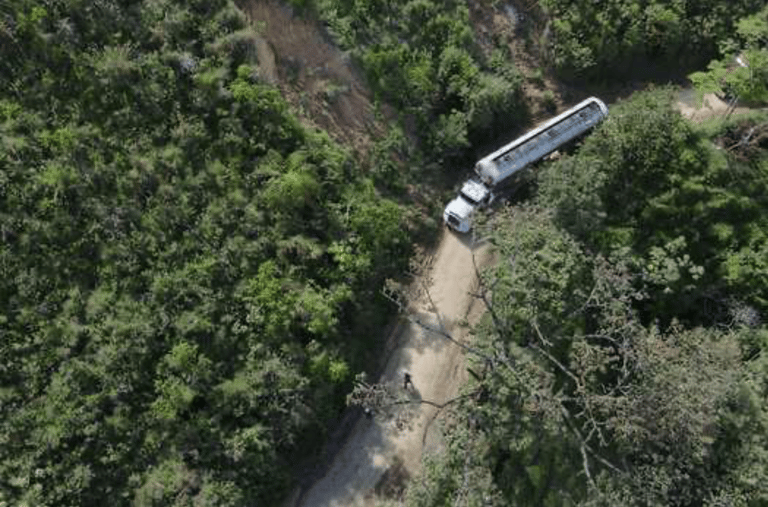 Aerial view of a white fuel tanker truck driving on a narrow dirt road through a dense tropical forest.