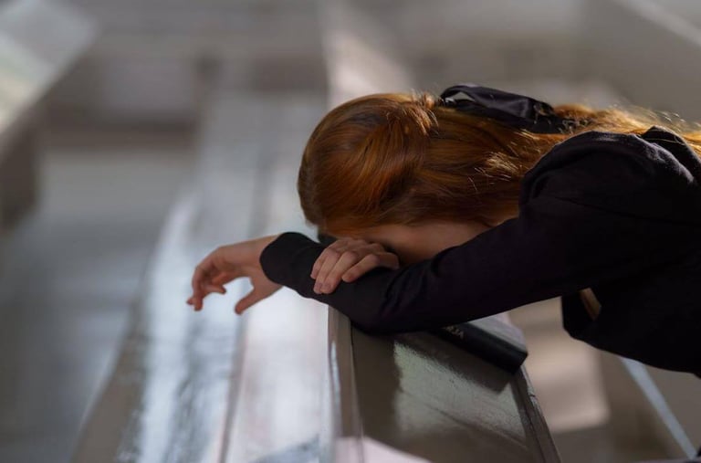 Individual resting head on crossed arms in a church pew, beside a book, in quiet reflection.