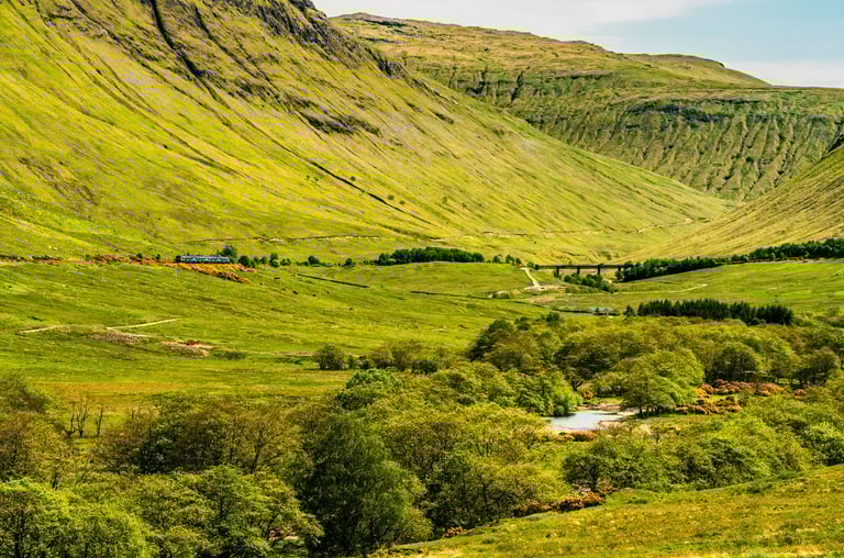 Vallée verdoyante de Glencoe en Écosse, bordée de montagnes majestueuses sous un ciel lumineux et dégagé.