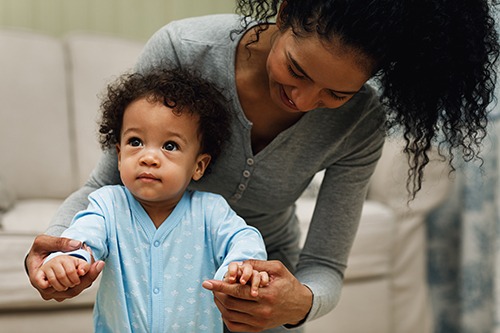 woman bent over holding hands of toddler learning to walk
