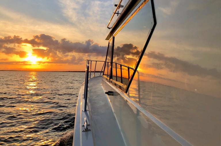 A luxury yacht cruising on the ocean at sunset with orange clouds reflecting on the water and cabin glass.