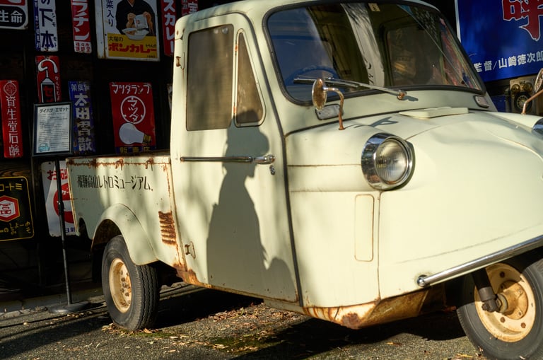 Vintage white Daihatsu Midget three-wheeled truck parked in front of retro Japanese metal signs.