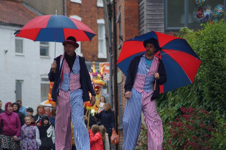 Two circus performers on stilts wearing striped costumes and holding red and blue umbrellas in a street parade.