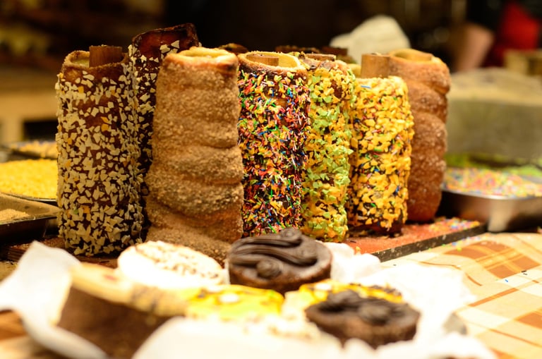 Freshly baked chimney cakes with assorted toppings like sprinkles and nuts at a pastry market stall.