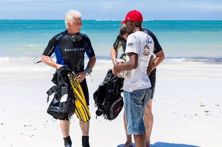 Scuba divers in wetsuits holding gear on a tropical white sand beach getting ready for a dive.