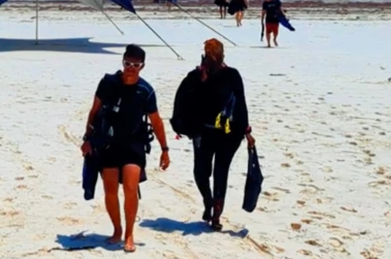 Scuba divers walking on a white sand tropical beach carrying diving gear near a blue ocean.