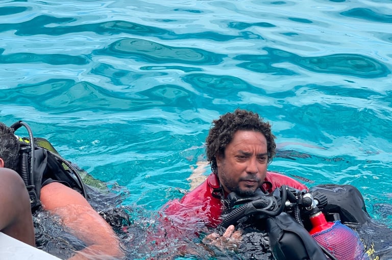Scuba diver wearing a red shirt and full dive gear floating in clear blue tropical ocean water.