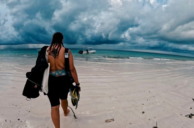 A scuba diver walks across Diani Beach's white sand beach toward a boat under dramatic storm clouds.