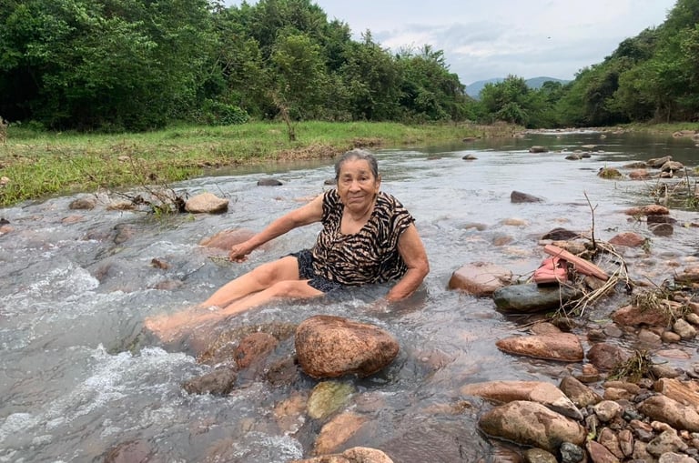 An elderly woman smiling while sitting in a shallow, rocky river surrounded by lush tropical greenery.