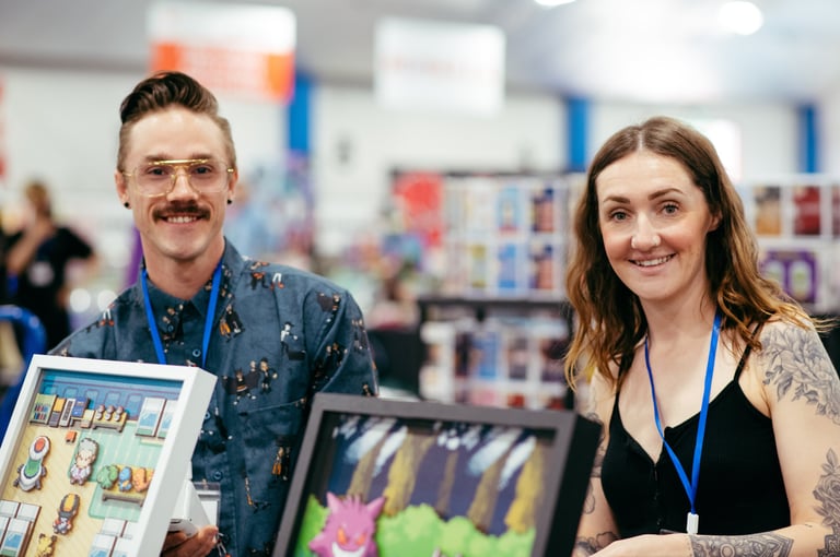 Two smiling vendors stand in front of the video game themed art they have for sale.