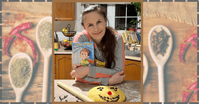 A woman smiling, holding a children's book titled "Living Dough," with a dough face on the table.