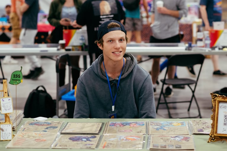 A smiling vendor sits at a table in front of his trading cards for sale.