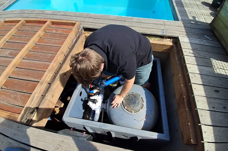 A technician performing pool maintenance on a sand filter system beneath a wooden deck.