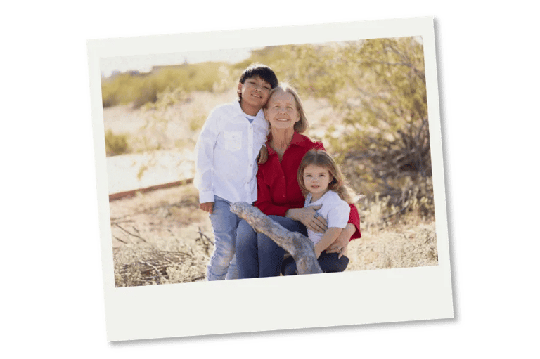 A grandmother smiling with her two young grandchildren in a sunny outdoor desert setting.