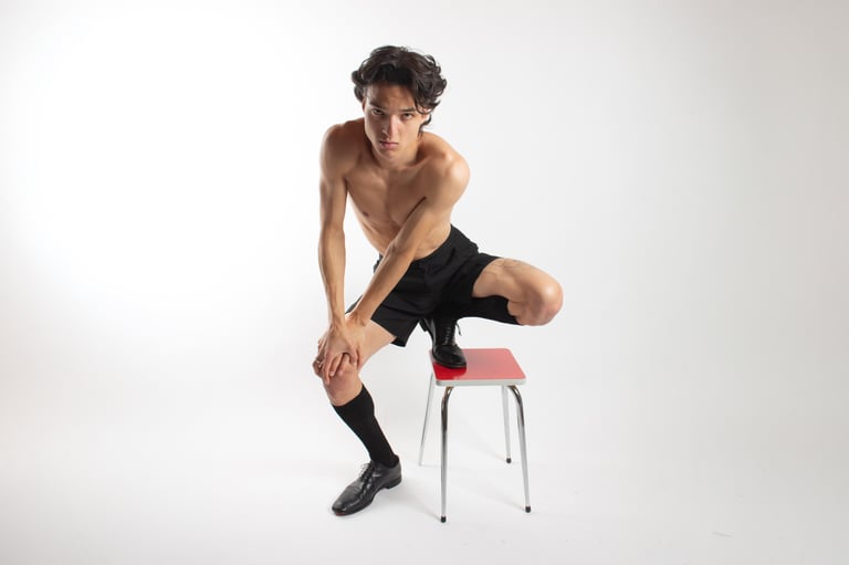 Shirtless male model in black shorts and dress shoes posing on a red stool in a studio.