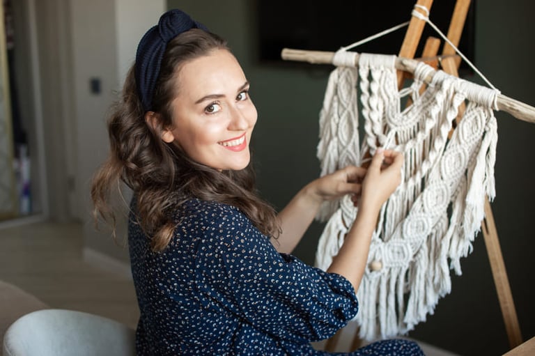 Mujer sonriente elaborando un tapiz de macramé blanco en un caballete de madera en casa.