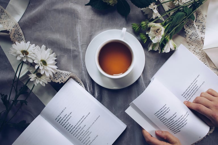 Flat lay of a person reading an open book next to a cup of hot tea and white flowers.