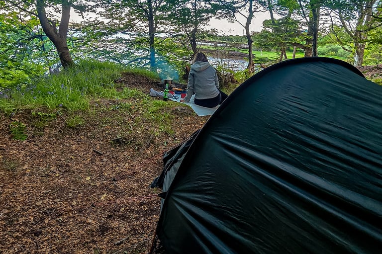 Campement en forêt en Écosse, avec une tente noire et une personne assise près d'un feu, au bord d'un lac.