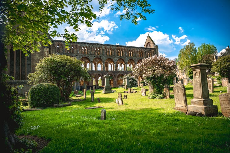 Ruines majestueuses de l'Abbaye de Jedburgh sous un ciel bleu, entourées d'arches gothiques, verdure et pierres tombales.
