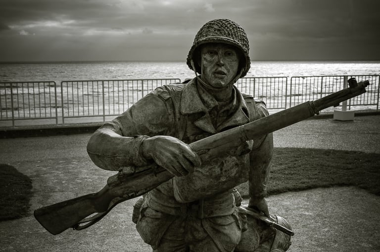 Statue en bronze d'un soldat courant avec un fusil, traînant un camarade tombé. Mer, obstacles et ciel nuageux en fond.