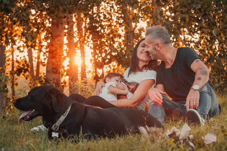 una coppia con un bambino in allattamento siede all'ombra di alberi al tramonto col proprio cane