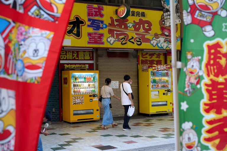 Two people walking past yellow vending machines on a colorful street in Osaka, Japan.
