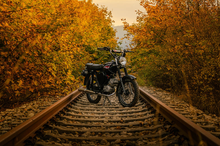Vintage black motorcycle parked on railroad tracks surrounded by golden autumn forest foliage.