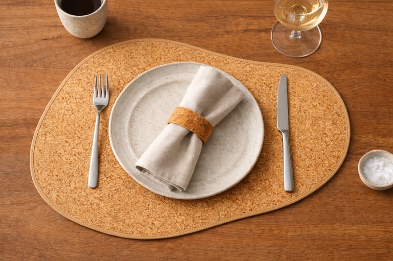 Minimalist table setting with a ceramic plate, linen napkin, and cork placemat on a wooden table.