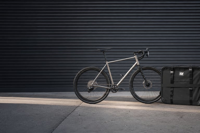 A titanium gravel bike leaning against a black travel bike bag in front of a dark shutter door.