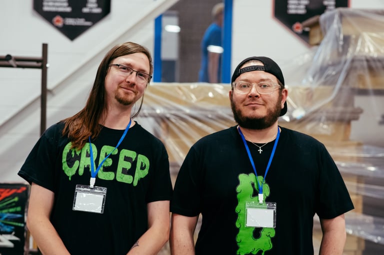 Two vendors with cool nerdy t-shirts stand smiling at their booth.