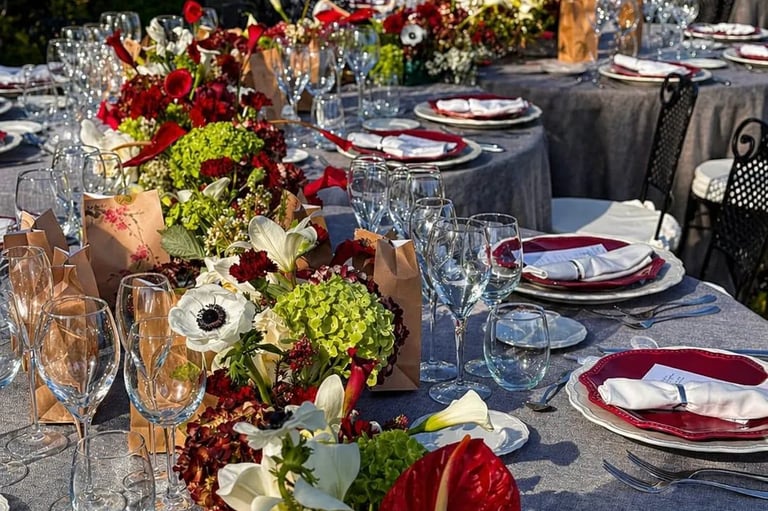 An elegantly set long outdoor dining table with a vibrant floral runner featuring red and white blossoms.
