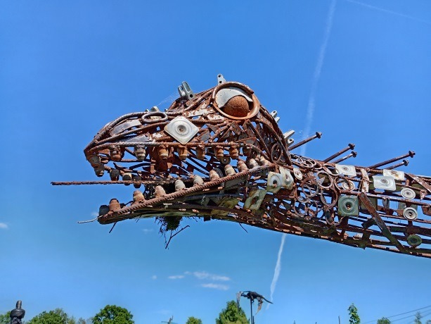A rustic metal dinosaur sculpture made from scrap metal and recycled parts against a blue sky.
