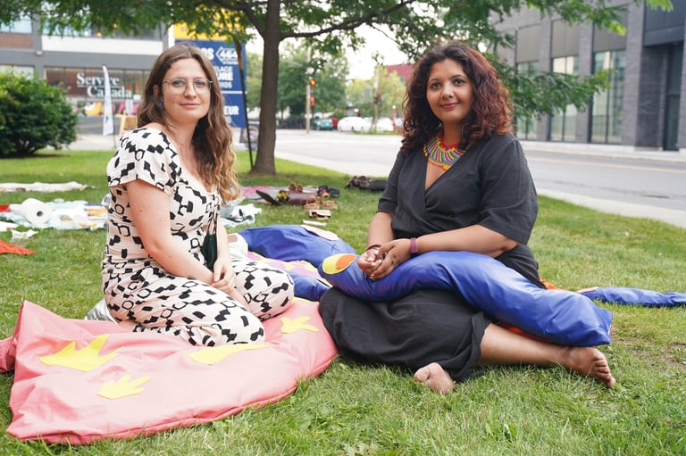 Two women pose for a portrait surrounded by soft body parts on the grass.